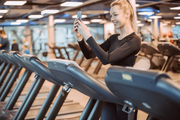 Fitness model listening music on smart phone charching positive energy before workout. Healthy fitness lifestyle concept with copy space. Athletic Sports Woman in Gym Puts on Wireless Headphones. - 写真・画像
