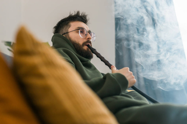 Millénaire barbu ou gen z homme fumant narguilé tout en se relaxant sur le canapé à la maison - temps de refroidissement et de repos - Photo, image