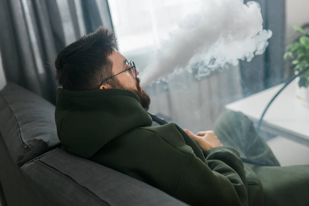 Millénaire barbu ou gen z homme fumant narguilé tout en se relaxant sur le canapé à la maison - temps de refroidissement et de repos - Photo, image