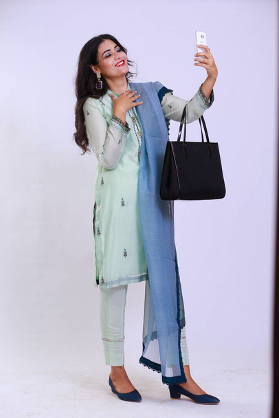 Mulher paquistanesa descendente, vestida com trajes tradicionais, vestindo um kamiz e shalwar. Ela tem o cabelo aberto e está usando um smartphone, bem como carregando uma bolsa.  - Foto, Imagem