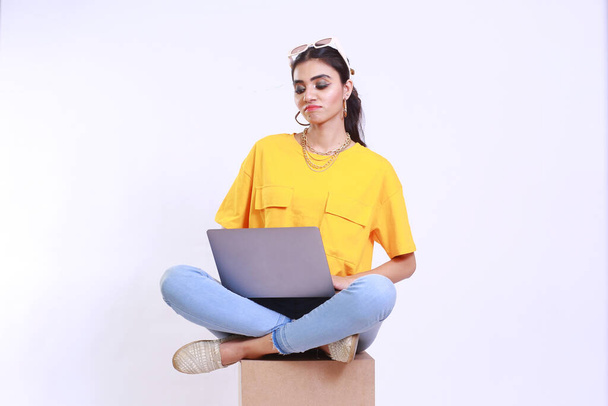 Young girl working online on a laptop computer sitting on a box stool showing un happy and un satisfied emotion, Fashionable trendy girl is wearing yellow tshirt. Isolated on a white studio background - 写真・画像