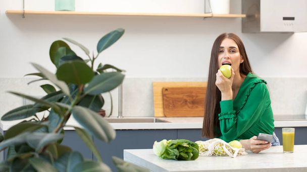 An attractive brunette holds a green apple standing at the kitchen table. Healthy lifestyle - Foto, immagini