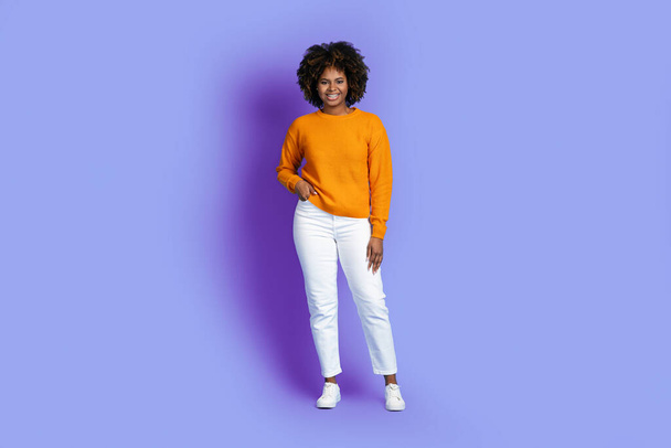 Happy cheerful beautiful young african american woman with bushy hair in bright stylish outfit posing alone on purple studio background, smiling at camera, full length shot - Fotografie, Obrázek