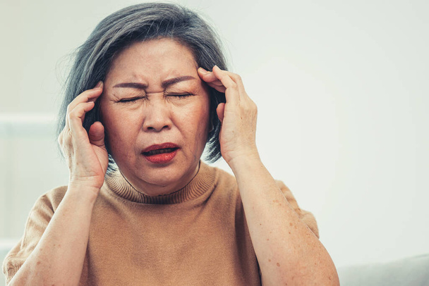 An agonizing senior woman with a headache, compress her temple with both hands at her contented living room. Senior health, nursing home, caretaker service. - Photo, Image