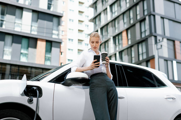 Focus businessman using phone, leaning on electric vehicle, holding coffee with blurred city residential condo buildings in background as progressive lifestyle by renewable and sustainable EV car. - 写真・画像