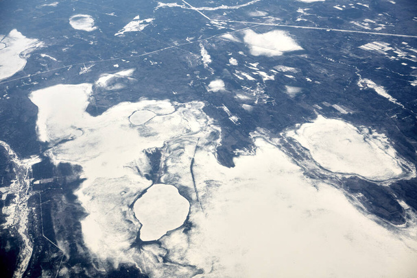 Aerial view from airplane window over clouds top to snow covered rivers, fields and roads, winter fresh frosty air. Beautiful hazed sky aerial view to Earth troposphere, snowy background texture - 写真・画像