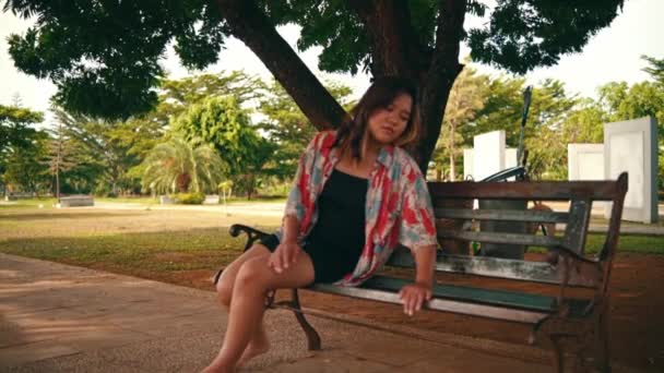 азиатка заснула на скамейке в парке перед большим тенистым деревом с очень усталым выражением лица в тот день - Кадры, видео