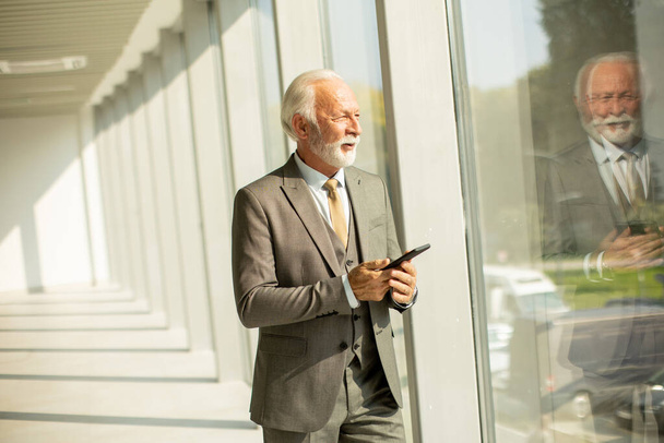 A senior business man stands in an office hallway, focused on his mobile phone. He is dressed in formal attire, exuding confidence and professionalism - Φωτογραφία, εικόνα