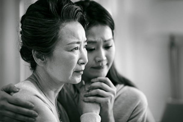 truchlící starší asijské matka a dospělá dcera držící se za ruce, černá a bílá - Fotografie, Obrázek