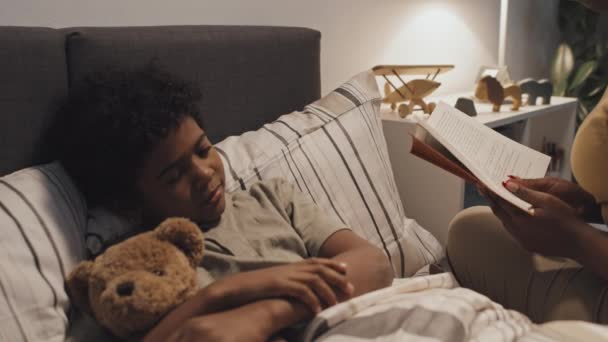 Yatağın kenarında oturan, elinde oyuncak ayıyla evde uyuyan sevimli siyahi çocuğa kitap okuyan bir ebeveyn. - Video, Çekim