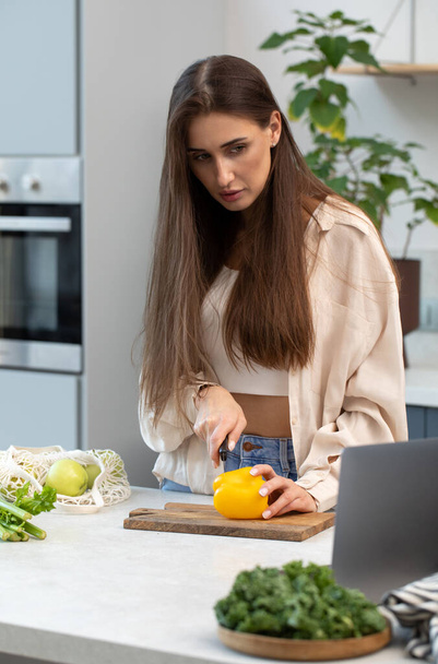 Attractive woman cuts vegetables for salad while looking at laptop screen. Video recipe. Healthy food. - Fotografie, Obrázek