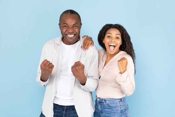 Hurra. Euphorisches afrikanisch-amerikanisches Paar ballt die Fäuste und ruft begeistert aus, Mann mittleren Alters und junge Frau feiern Erfolg, posieren vor blauem Hintergrund - Foto, Bild
