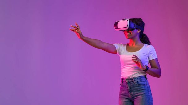Esperienza virtuale. Sorridente Black Lady toccando aria durante la riproduzione di videogiochi in VR Headset, Eccitato giovane donna afroamericana in piedi nella luce al neon su sfondo viola Studio, Panorama - Foto, immagini