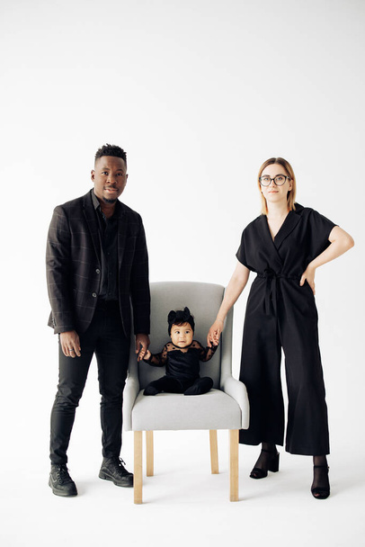 Happy interracial family stands near their baby daughter sitting in armchair. Concept of interracial family and unity between different human races. - Фото, изображение