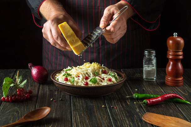 Повар добавляет твердый сыр к спагетти со специями и травами в тарелке. Рабочая обстановка на кухне ресторана - Фото, изображение
