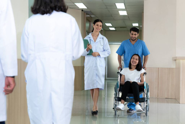 Lekarz i pielęgniarz przewożą pacjentkę na wózku inwalidzkim sterylnym szpitalnym korytarzem. Opieka zdrowotna i opieka pielęgniarska nad niepełnosprawnym pacjentem w koncepcji szpitala. - Zdjęcie, obraz