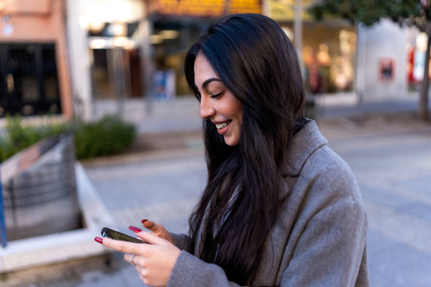 Optimista fiatal nő hosszú sötét hajjal meleg kabátot visel mosolygós és üzenetküldő mobiltelefon, miközben séta az utcán a városban - Fotó, kép