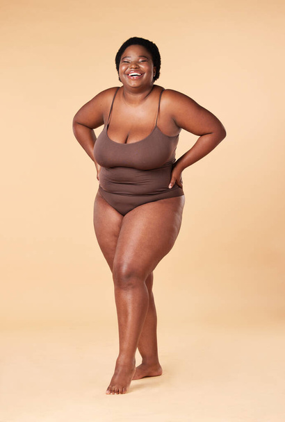 Portrait, happy or plus size with a model black woman posing hands on hips in studio on a beige background. Wellness, underwear and smile with a female standing proud or confident for body positivity. - Fotó, kép