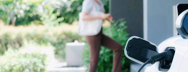 Focus EV charger plugged into EV car at home charging station with blurred background of progressive woman walking in background. Elective vehicle powered by clean energy for eco-friendly concept. - 写真・画像