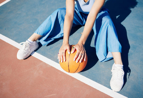 Basketball, hands and woman on the outdoor court floor with urban fashion for influencer beauty. Sport, model and young person sitting with gen z clothes with sports game equipment in summer. - 写真・画像