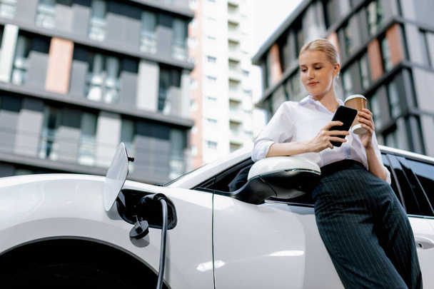 Focus businessman using phone, leaning on electric vehicle, holding coffee with blurred city residential condo buildings in background as progressive lifestyle by renewable and sustainable EV car. - Foto, Imagen