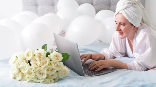 Woman in white clothes and towel on her head lying on a bed with white color balloons and bouquet of roses. Woman works at home, using her laptop, computer. Birthday, wedding. Copy space. Preparation. - Felvétel, videó