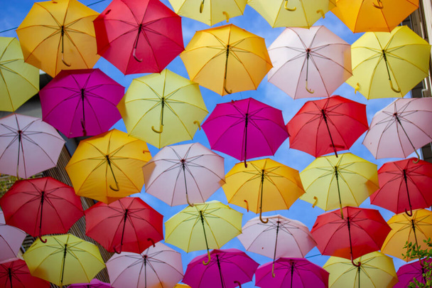 Φόντο από πολλά πολύχρωμα φωτεινά χρωματιστές ομπρέλες σε μια μπλε θέα στον ουρανό από κάτω. Γιορτινή διακόσμηση των δρόμων της πόλης. Σχέδιο ταξιδιού και αναψυχής. Πολύχρωμες ομπρέλες κρέμονται. - Φωτογραφία, εικόνα