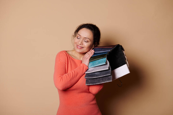 Attractive smiling woman, interior designer, decorator or customer choosing upholstery materials, leaning her head on fabric swatches, posing with her eyes closed on beige background. Home improvement - Zdjęcie, obraz