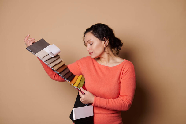 Multi-ethnic pretty woman touching and testing fabric samples of upholstery materials, isolated on beige color background. Interior design and home improvement concept. People and Creative profession - Photo, Image