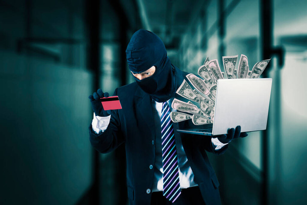 Hacker wearing business suit and black mask and holding laptop stealing money from a credit card - Photo, image
