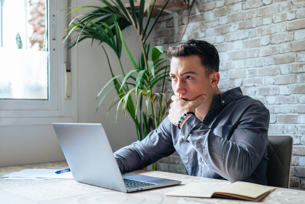 Thoughtful businessman touching chin, pondering ideas or strategy, sitting at wooden work desk with laptop, freelancer working on online project, student preparing for exam at home - Foto, imagen