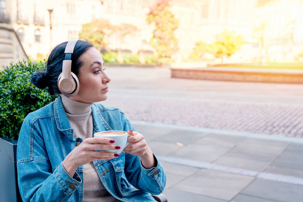 woman in denim coat and headphones drinking coffee and listening music or  audiobook in the cafe. Lifestyle concept - Photo, Image