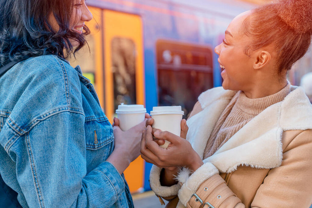 dos mujeres en una chaqueta de mezclilla está hablando entre sí, bebiendo café y esperando un tranvía en la parada Lifestyle foto - Foto, imagen