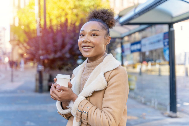 Веселая африканская женщина держит чашку кофе. Улыбающаяся брюнетка в коричневом свитере ждет трамвай. - Фото, изображение