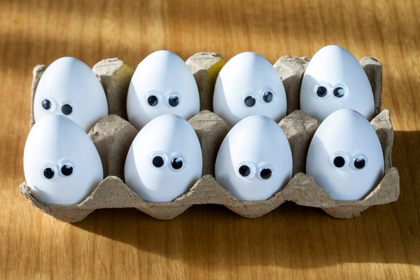 Funny faces on white eggs in carton box with organic chicken eggs on kitchen table closeup big animation eyes. humor, food and easter holiday concept. close up - Фото, изображение