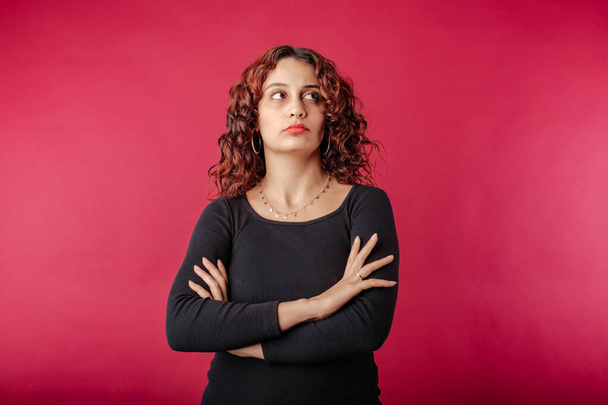 Portrait de jeune femme rousse portant une robe noire debout isolé sur fond rouge avec les bras croisés regarde l'espace de copie vide avec une expression de désapprobation. - Photo, image