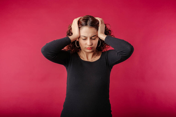 Красивая рыжая женщина в черном платье, стоящая изолированно на красном фоне рука на голове, головная боль, потому что стресс. Страдания от мигрени. - Фото, изображение