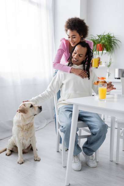 Улыбающаяся африканская американка обнимает бойфренда рядом с лабрадором и завтракает на кухне  - Фото, изображение