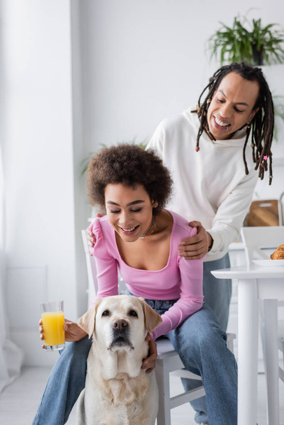 Mujer afroamericana positiva sosteniendo jugo de naranja cerca de labrador y novio en cocina  - Foto, Imagen