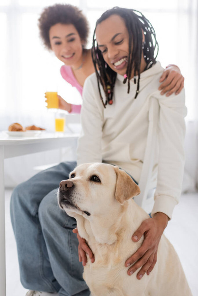 Labrador sentado cerca borrosa pareja afroamericana con desayuno en la cocina  - Foto, imagen