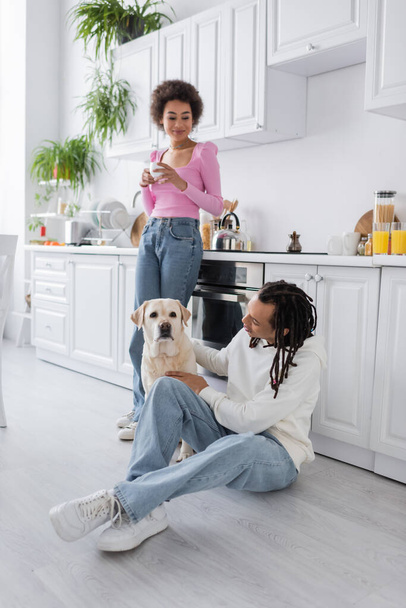 African american holding coffee near boyfriend petting labrador in kitchen  - Foto, afbeelding