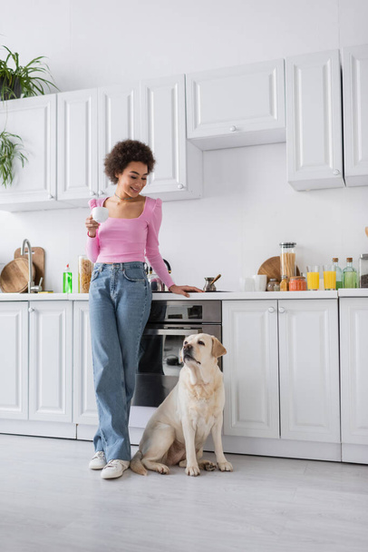 Positieve Afrikaanse Amerikaanse vrouw op zoek naar labrador terwijl het houden van koffie in de keuken  - Foto, afbeelding