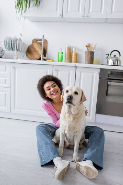 Positive Afroamerikanerin schaut Labrador in Küche an  - Foto, Bild