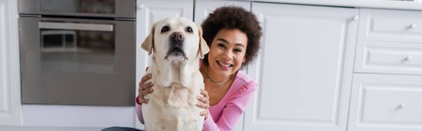 Happy african american woman petting labrador in kitchen, banner  - Photo, Image