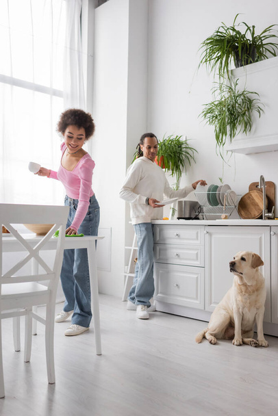 Positive african american woman holding cup and cleaning table near boyfriend and labrador in kitchen  - Photo, Image