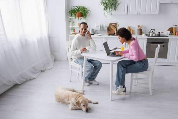 African american couple using laptop near drinks and labrador in kitchen  - Foto, Imagen