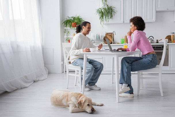 Cheerful african american couple using devices near drinks and labrador at home  - Fotoğraf, Görsel