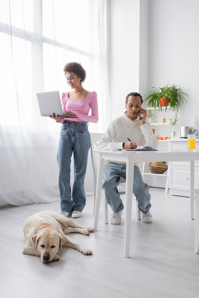 Mulher afro-americana positiva usando laptop perto namorado falando no smartphone e labrador em casa  - Foto, Imagem