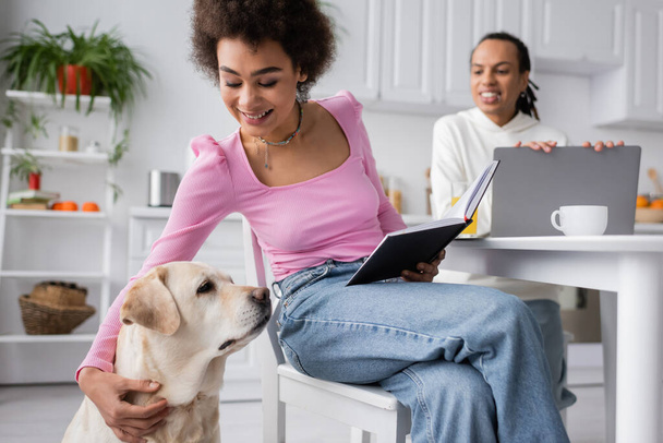 Cheerful african american woman holding notebook and hugging labrador near boyfriend in kitchen  - Foto, Imagem