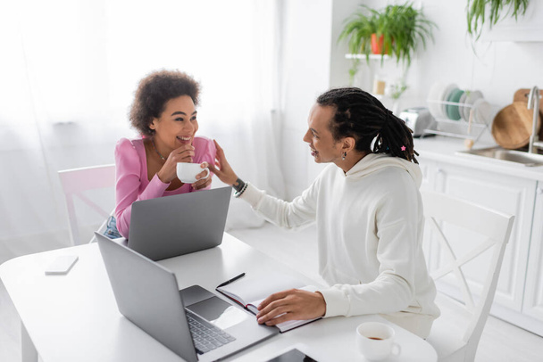 Cheerful african american couple talking near devices and coffee in kitchen  - Fotoğraf, Görsel